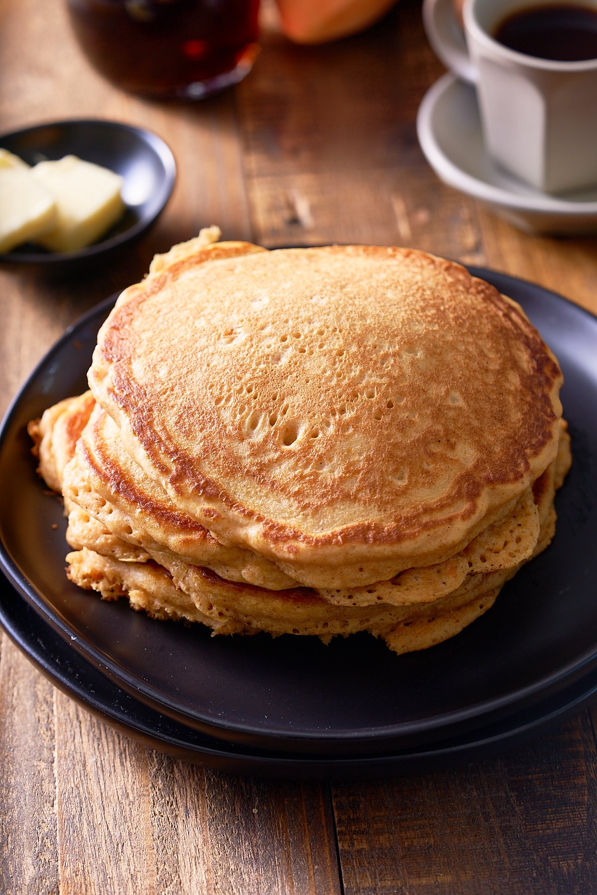 A plate topped with 3 sweet potato pancakes, with a cup of coffee and small dish of butter set alongside.