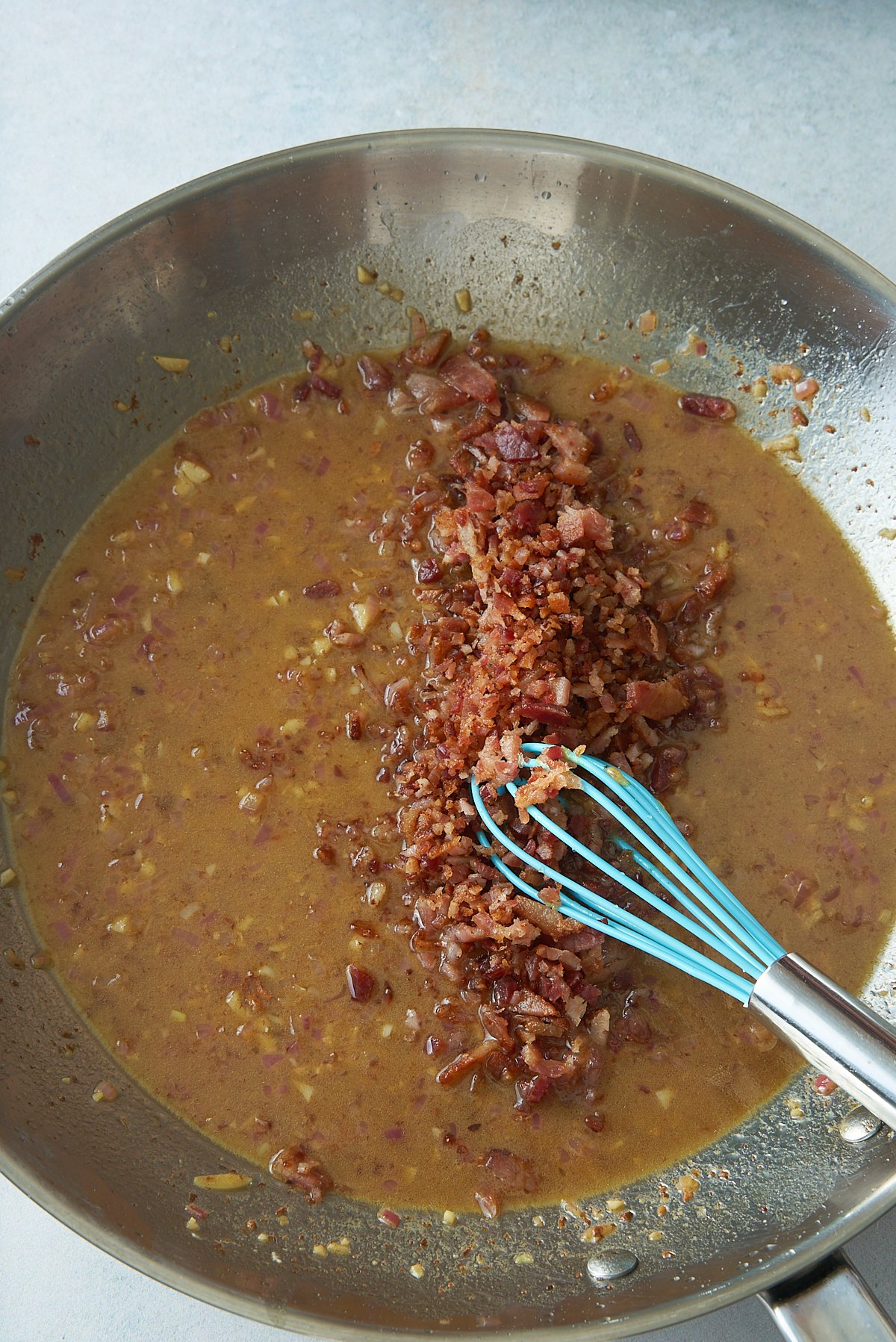 A large skillet of warm vinaigrette with chopped bacon being whisked in.