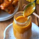 A glass jar filled with Carolina Gold BBQ sauce, with a spoon of sauce dripping into the jar and a dish of grilled chicken set alongside.