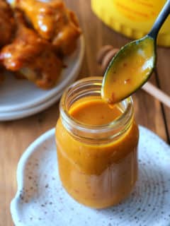 A glass jar filled with Carolina Gold BBQ sauce, with a spoon of sauce dripping into the jar and a dish of grilled chicken set alongside.