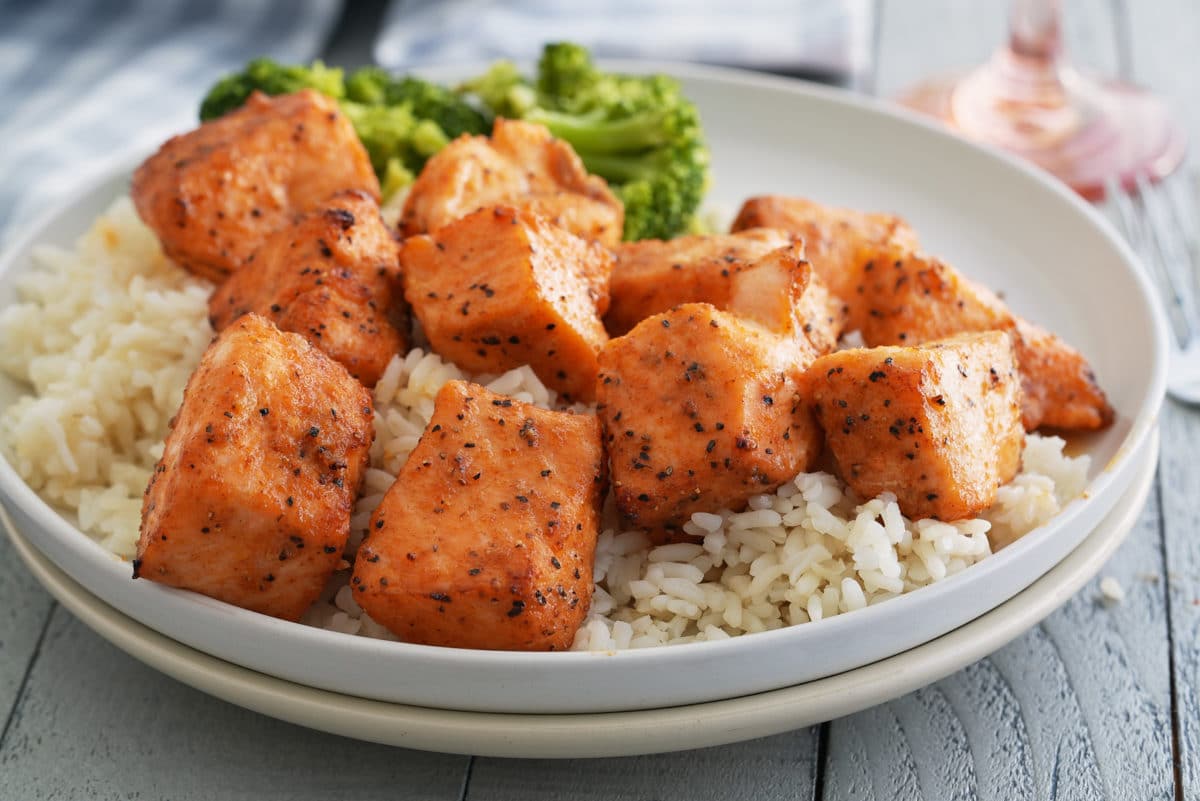 A white bowl of steamed white rice topped with salmon bites and a side of steamed broccoli.
