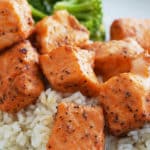 A white bowl of steamed white rice topped with salmon bites and a side of steamed broccoli.