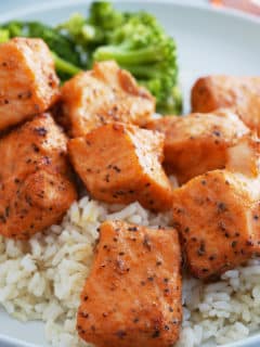 A white bowl of steamed white rice topped with salmon bites and a side of steamed broccoli.