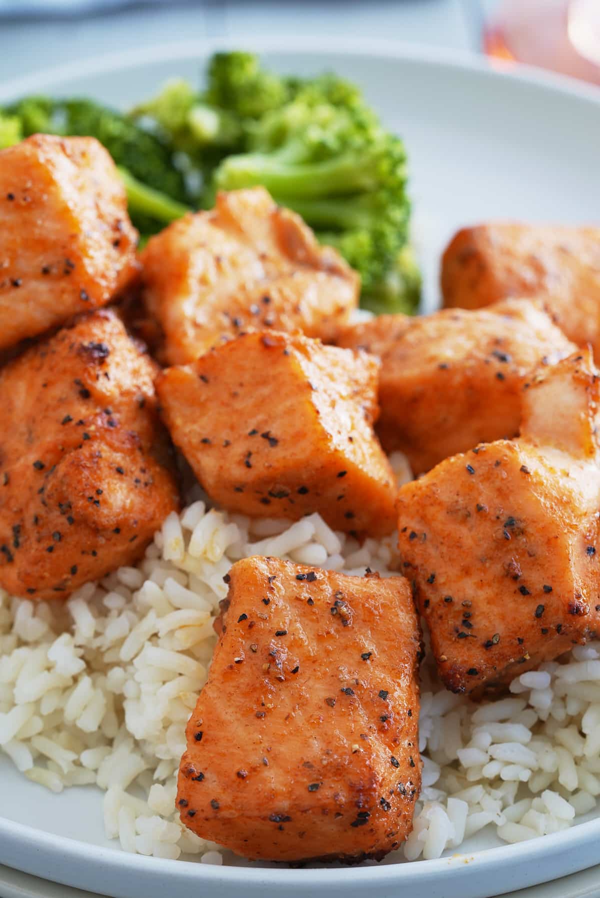 A white bowl of steamed white rice topped with salmon bites and a side of steamed broccoli.