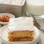 A white plate with a slice of glazed honey bun cake with honey, the whole cake in the tin and a glass of milk set alongside.