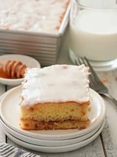 A white plate with a slice of glazed honey bun cake with honey, the whole cake in the tin and a glass of milk set alongside.