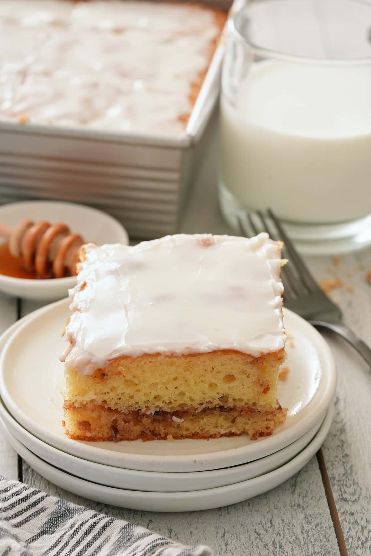 A white plate with a slice of glazed honey bun cake with honey, the whole cake in the tin and a glass of milk set alongside.