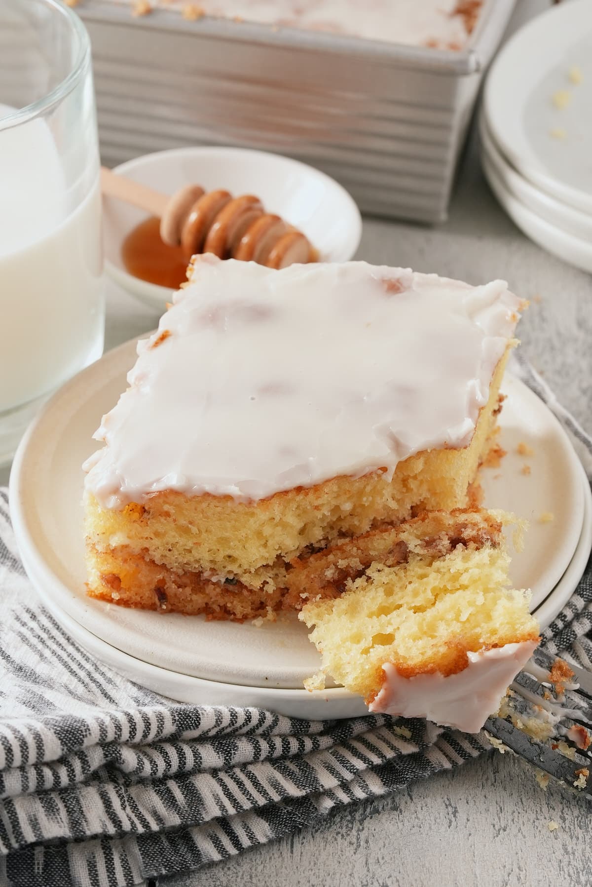 A white plate with a slice of glazed honey bun cake with honey, the whole cake in the tin and a glass of milk set alongside.