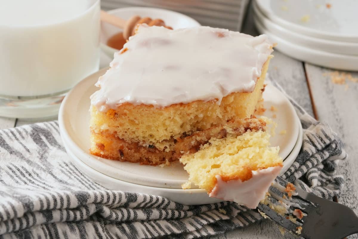 A white plate with a slice of glazed honey bun cake with honey and a glass of milk set alongside.