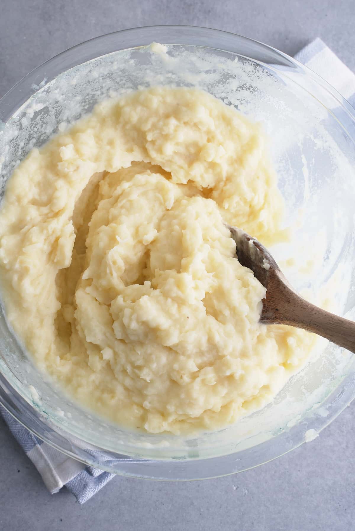A glass bowl filled with mashed potatoes being stirred with a wooden spoon.