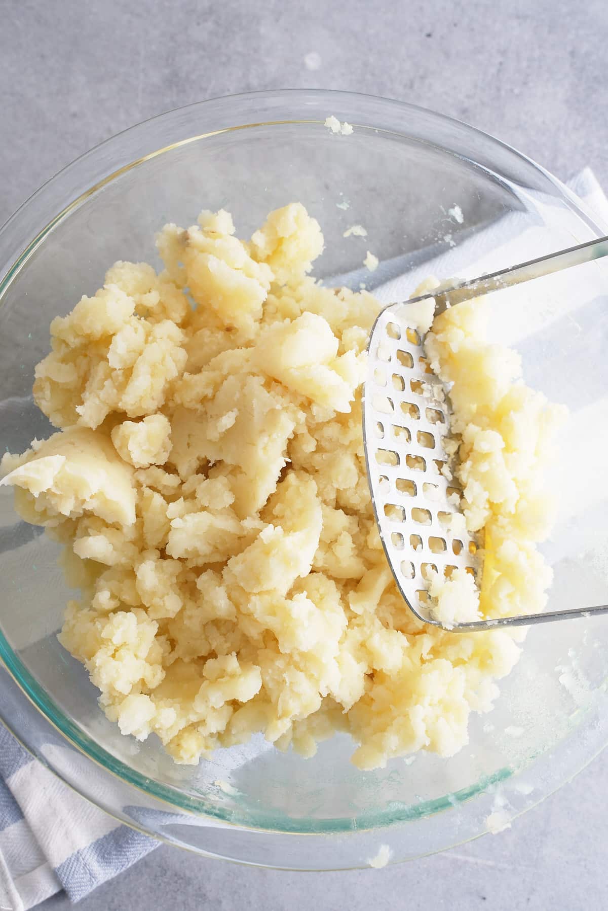 A glass bowl filled with cooked potato being mashed with a potato masher.