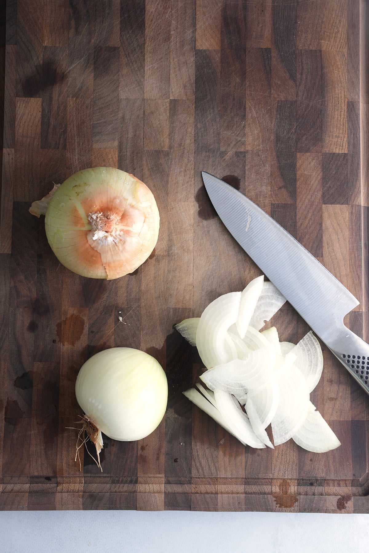 A wooden cutting board with a whole onion and a second onion being sliced with a sharp knife.