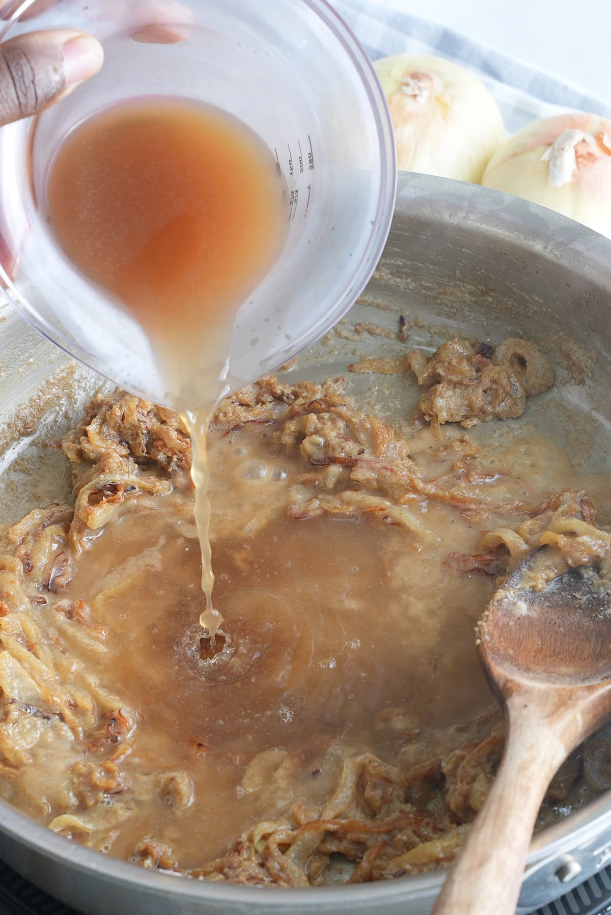 A jug of beef broth being stirred into a mixture of cooked onions and flour.