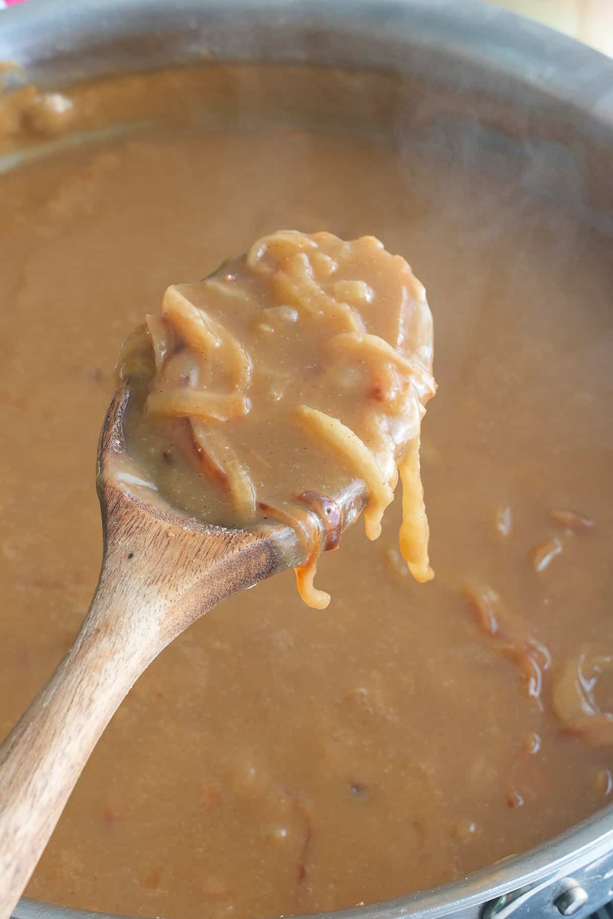 A large skillet of onion gravy with a wooden spoon of cooked sliced onions.