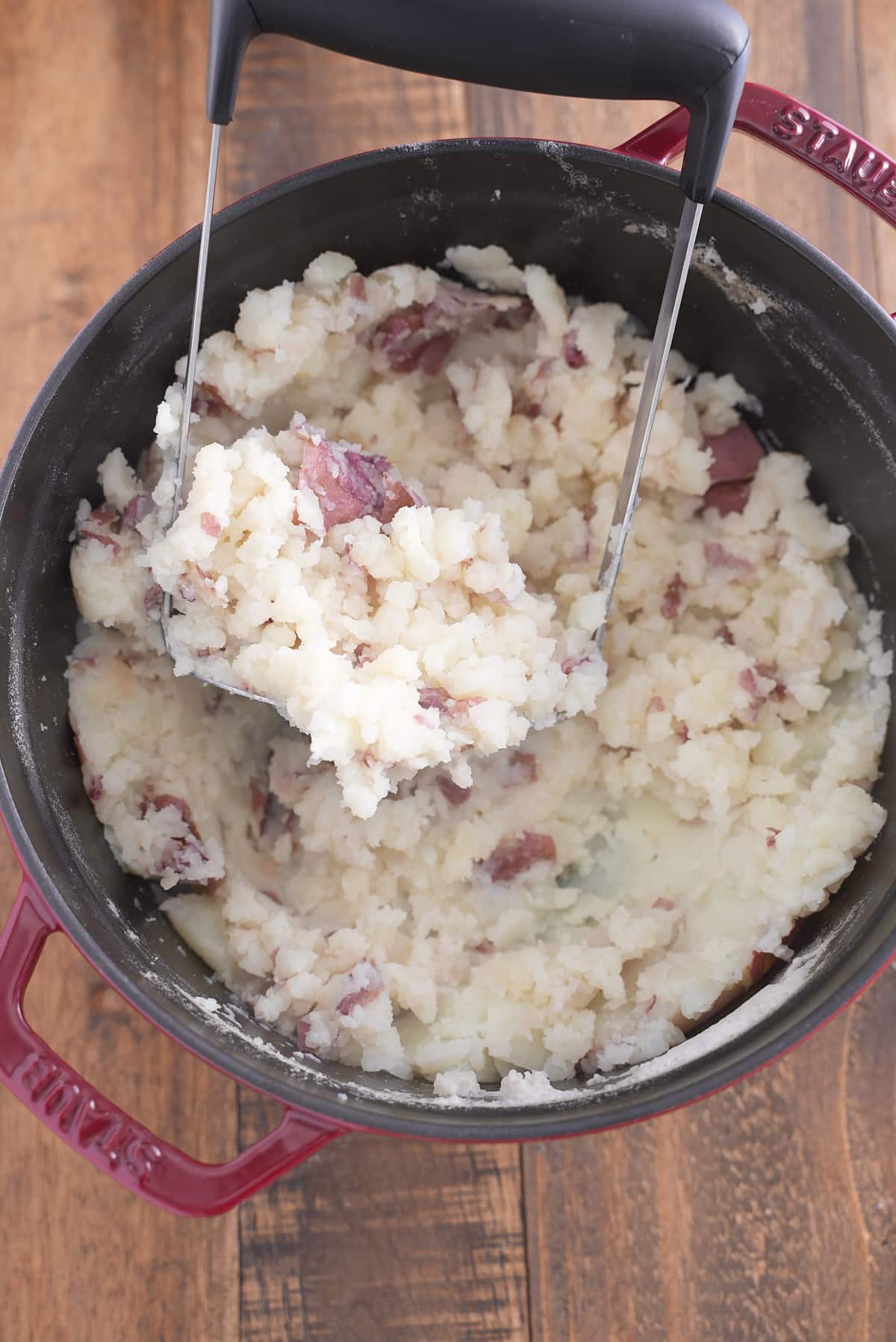 A Dutch oven filled with cooked red skinned potatoes being mashed with a potato masher.