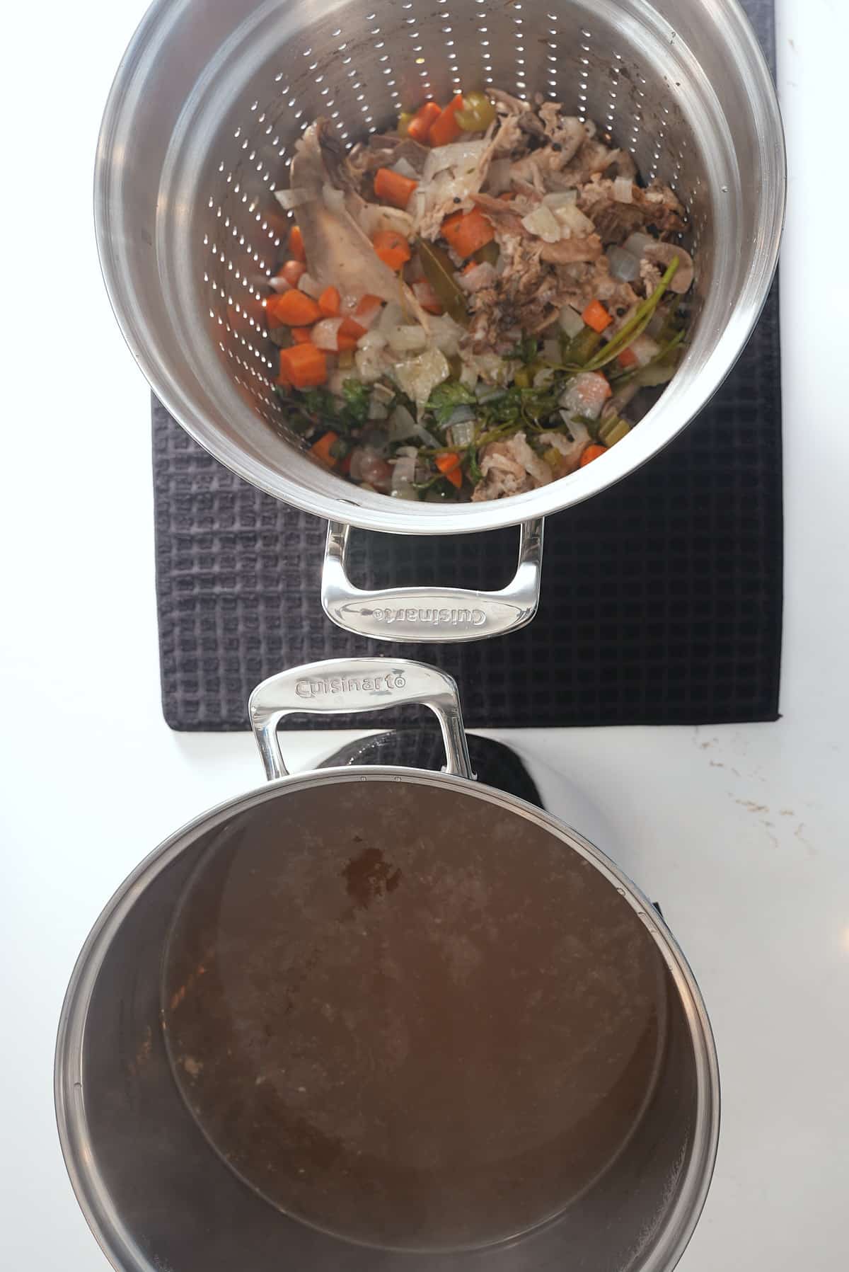 A large pot of strained turkey stock, with a colander of used vegetables and herbs set alongside.