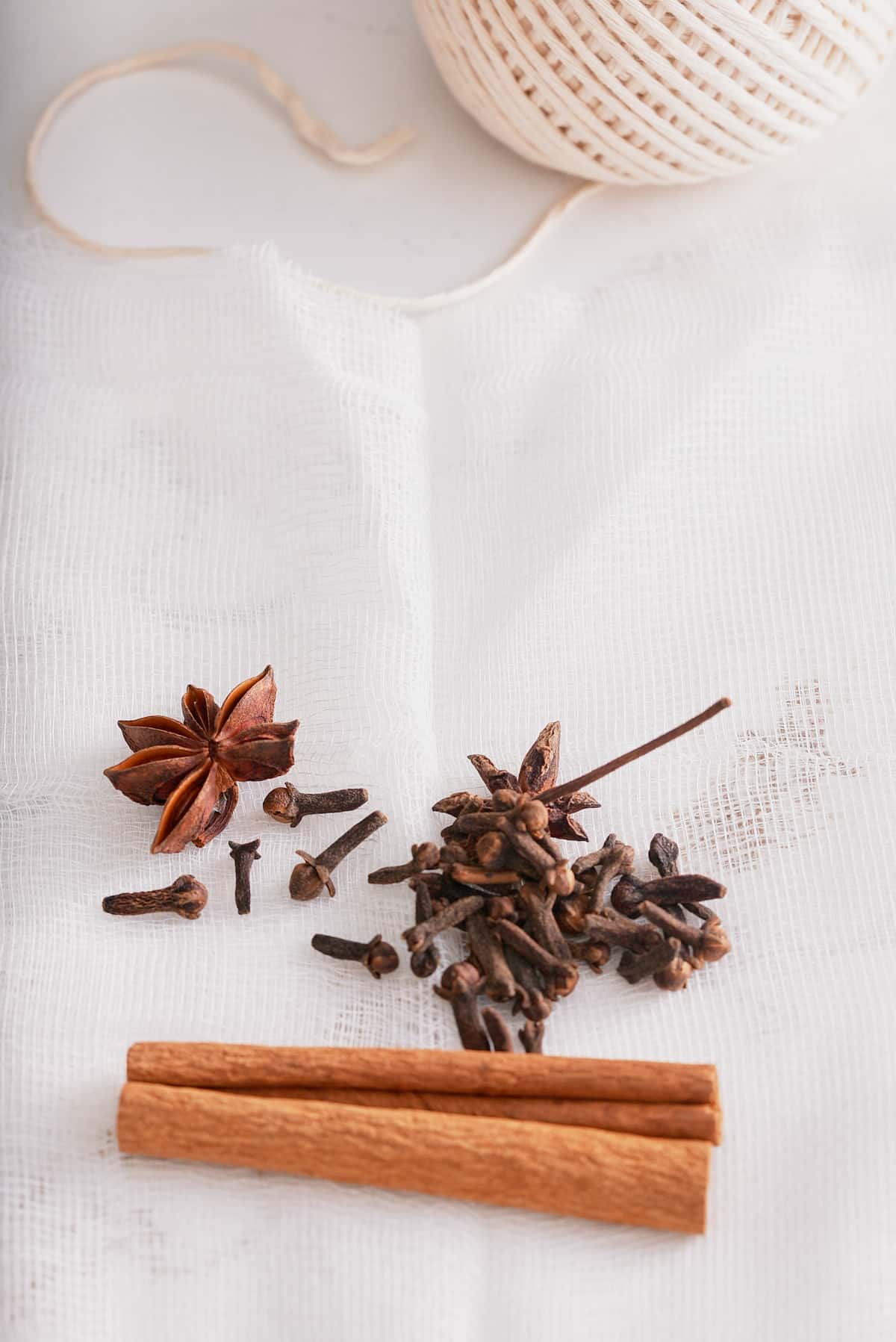 A cinnamon stick, whole cloves and whole star anise set of a piece of white cheesecloth.