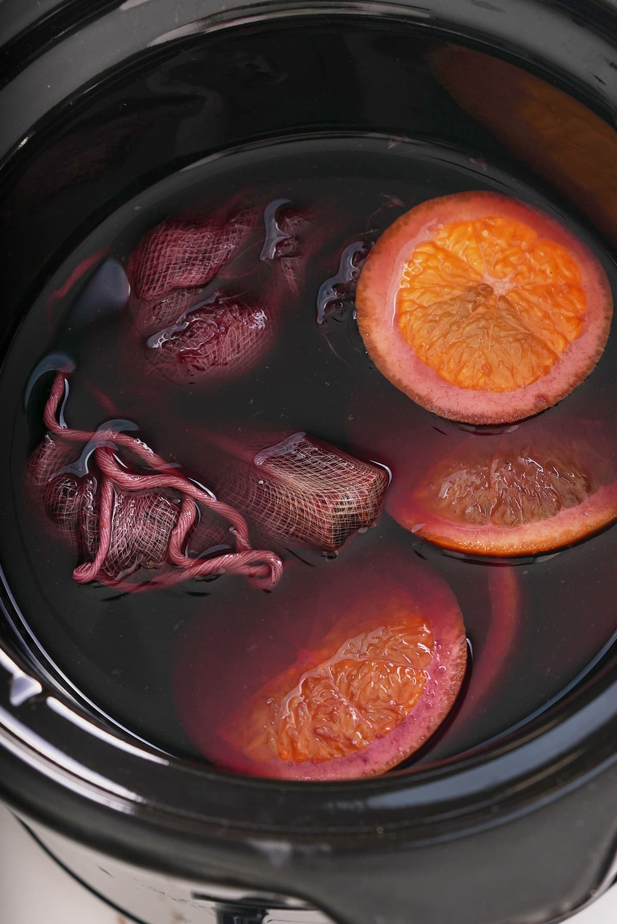 Slow cooker filled with red white, spice sachet and slices of fresh orange.