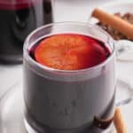 Two glass cups filled with mulled wine and topped with a slice of mulled orange with whole spices set alongside.