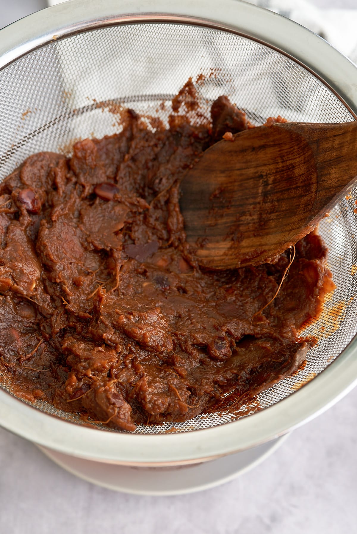 A wooden spoon bruising cooked tamarind pods through a sieve to release the water.