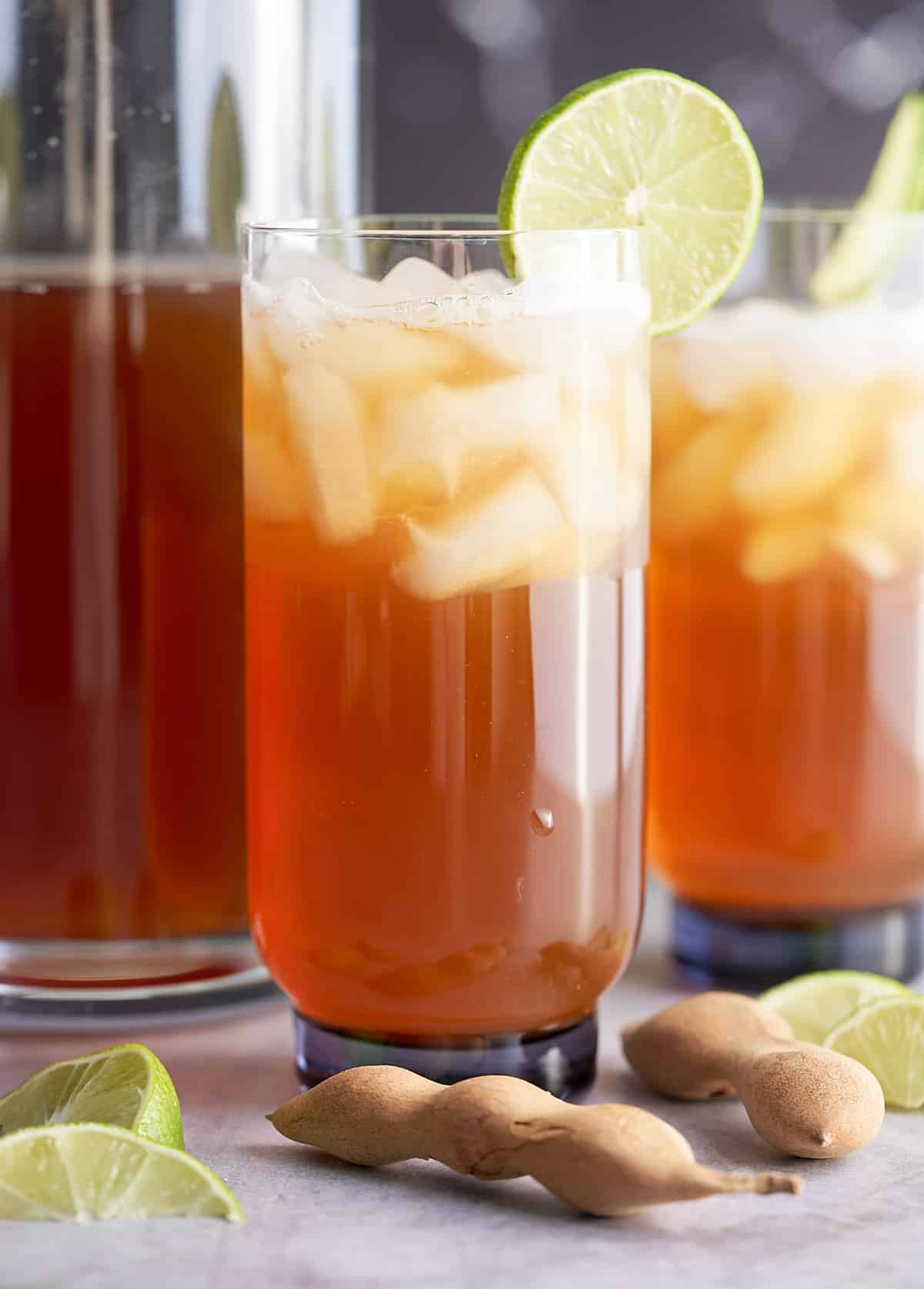 Two glasses of iced tamarind juice garnished with a slice of fresh lime with a pitcher of tamarind juice set alongside.