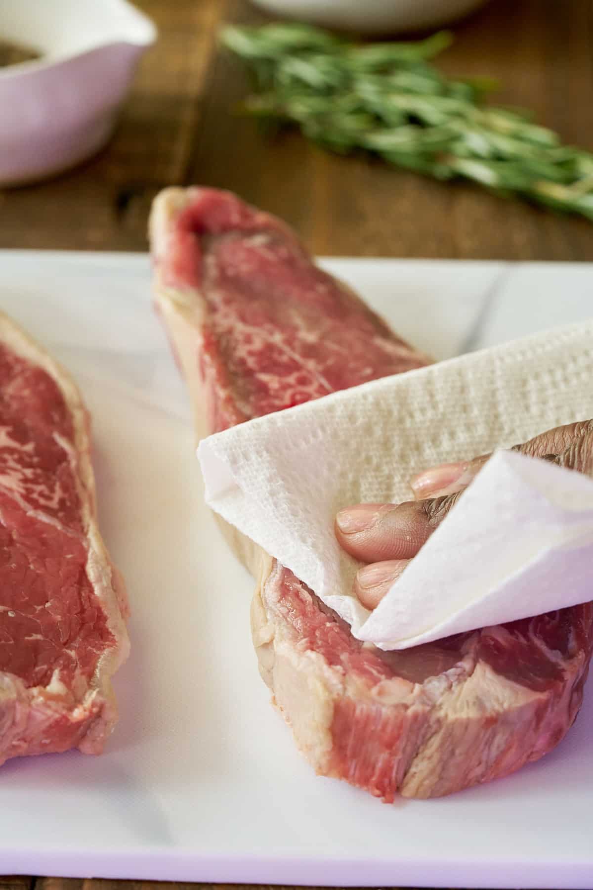 Two sirloin steaks on a cutting board being patted dry with kitchen paper.