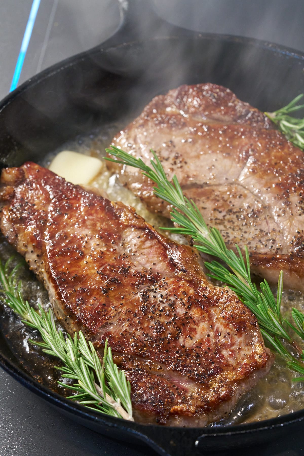 Two seared sirloin steaks in a cast iron skillet with added rosemary and butter.