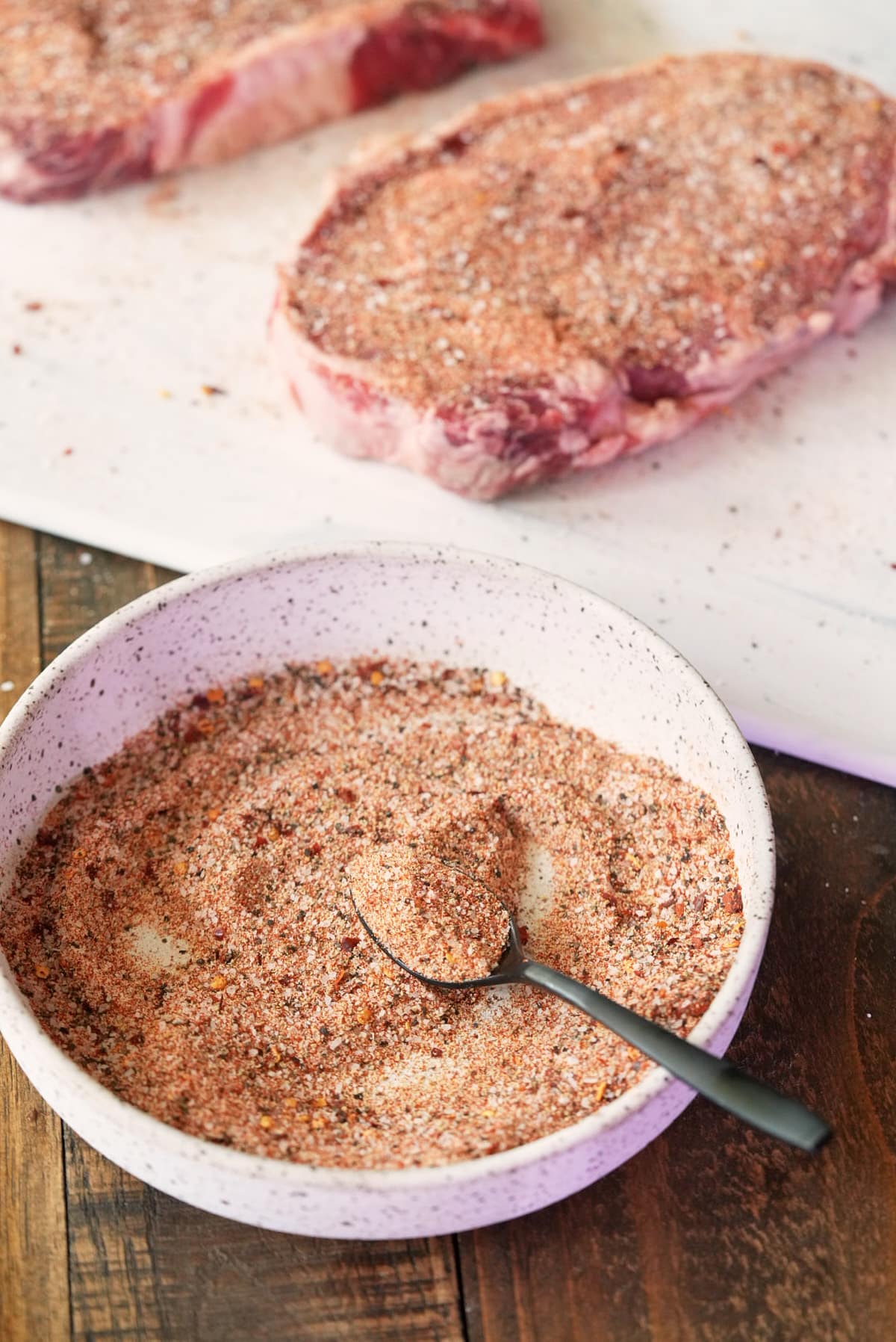 Steaks covered in homemade steak seasoning with a bowl of steak seasoning set alongside.