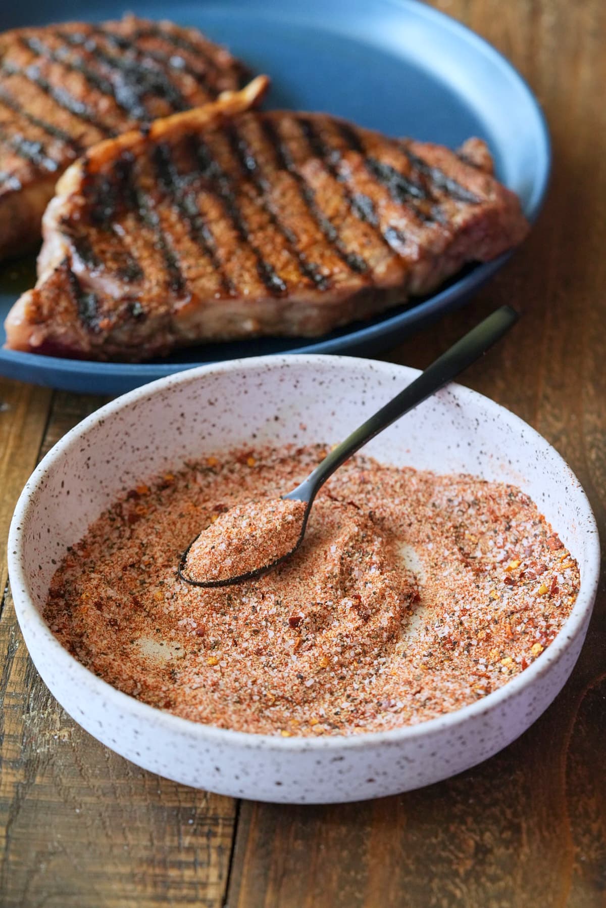 Two cooked steaks coated in steak seasoning with a bowl of steak seasoning set alongside.