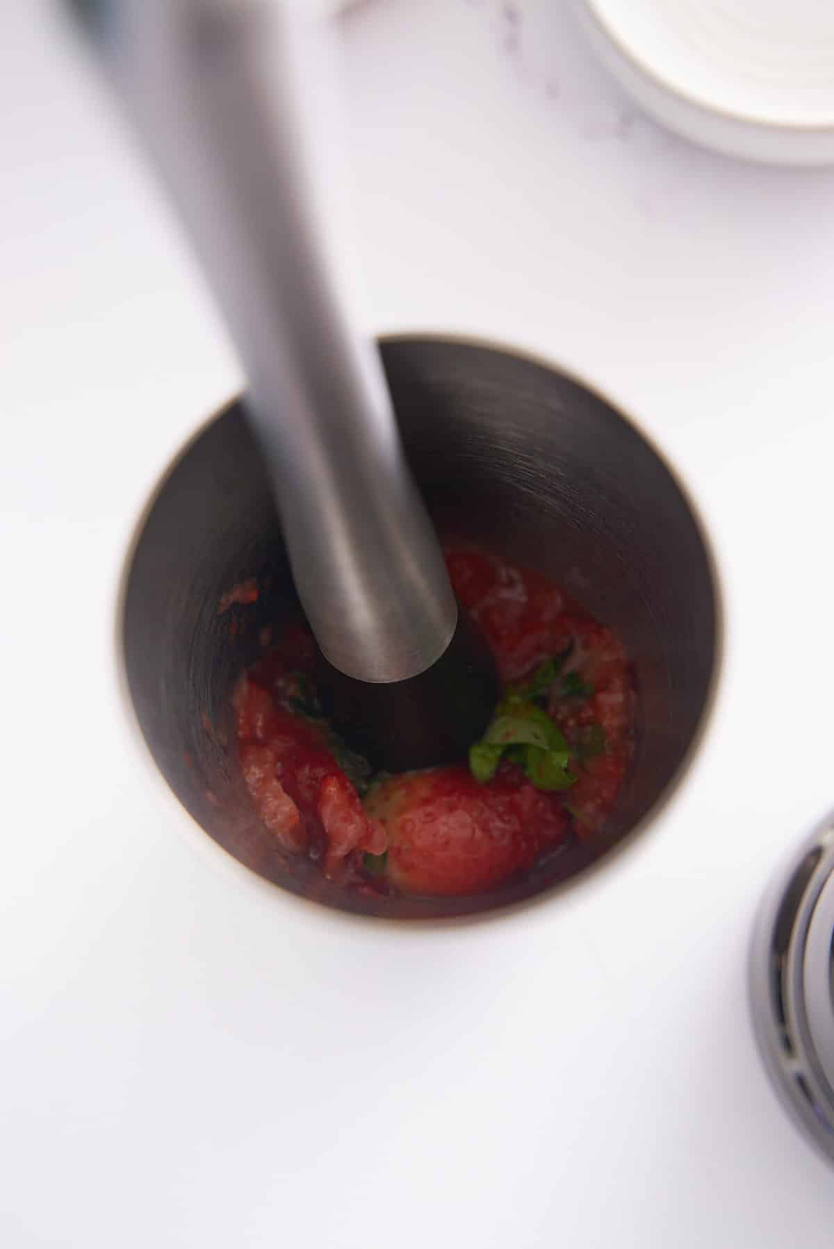 Strawberries and basil leaves being muddled and broken down in a cocktail shaker.