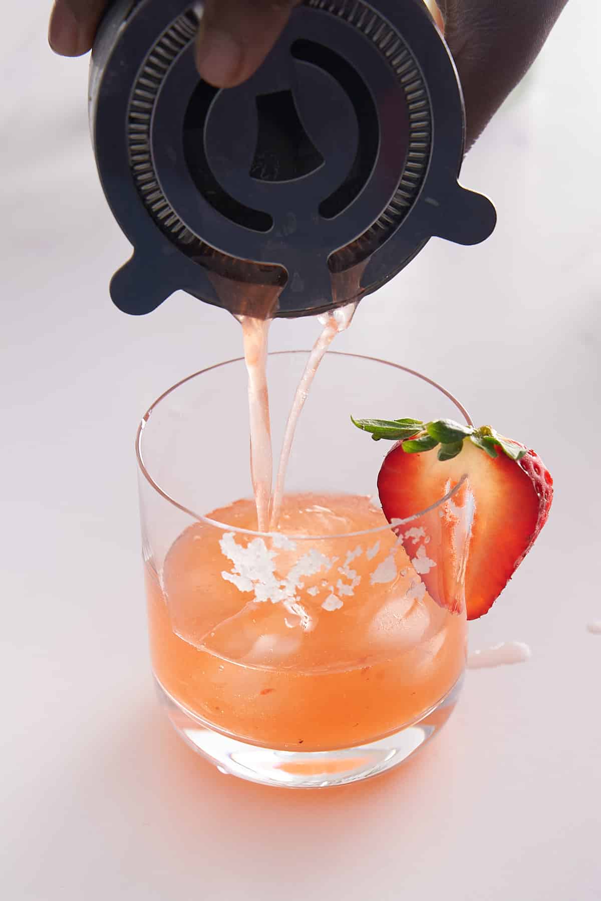 Strawberry basil margarita cocktail being poured from a cocktail shaker into a glass filled with icecubes.