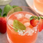 A glass filled with strawberry basil margarita served with ice and garnished with a slice of strawberry and a basil leaf.