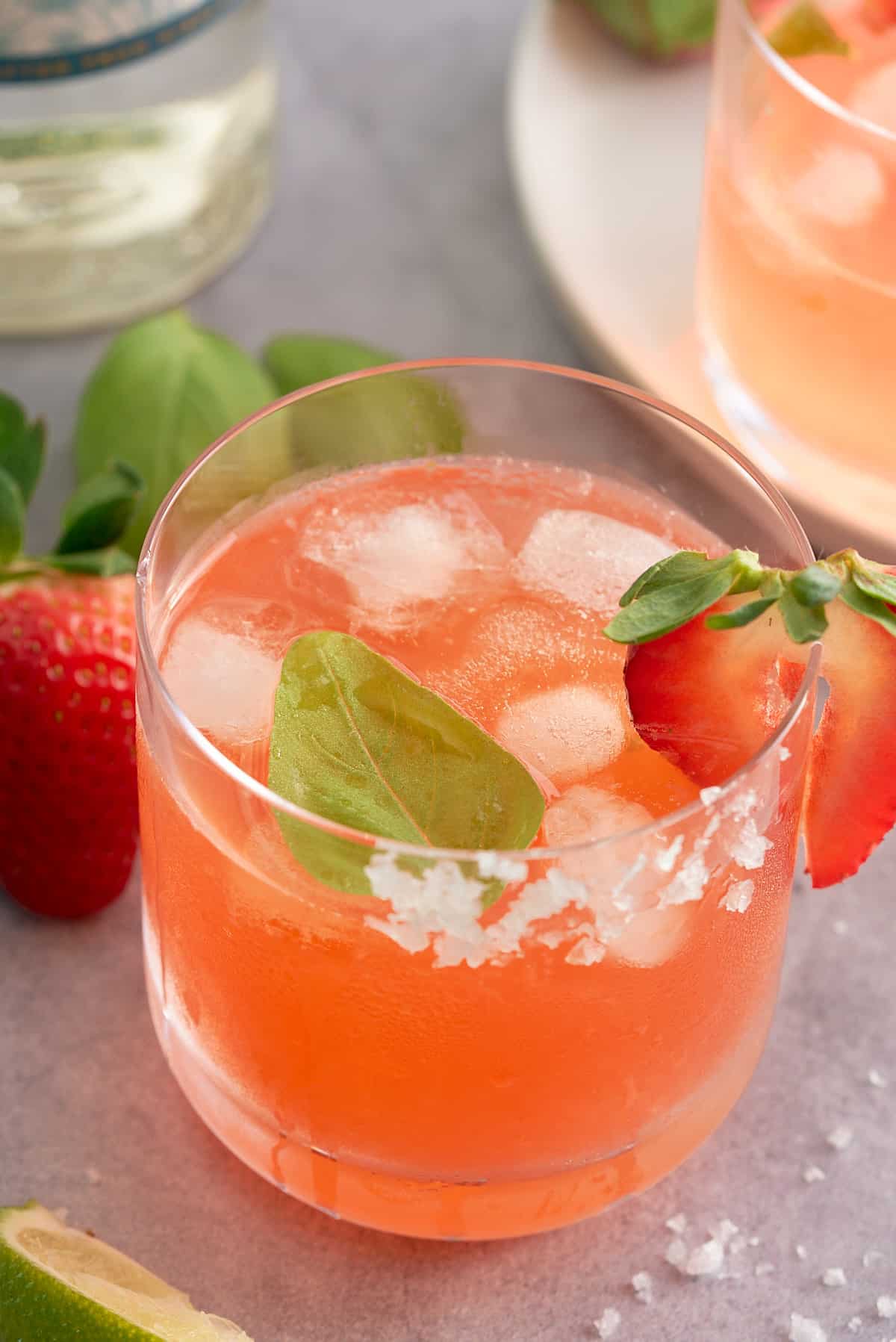 A glass filled with strawberry basil margarita served with ice and garnished with a slice of strawberry and a basil leaf.