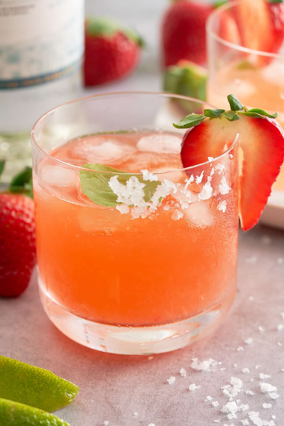A glass filled with strawberry basil margarita served with ice and garnished with a slice of strawberry and a basil leaf.