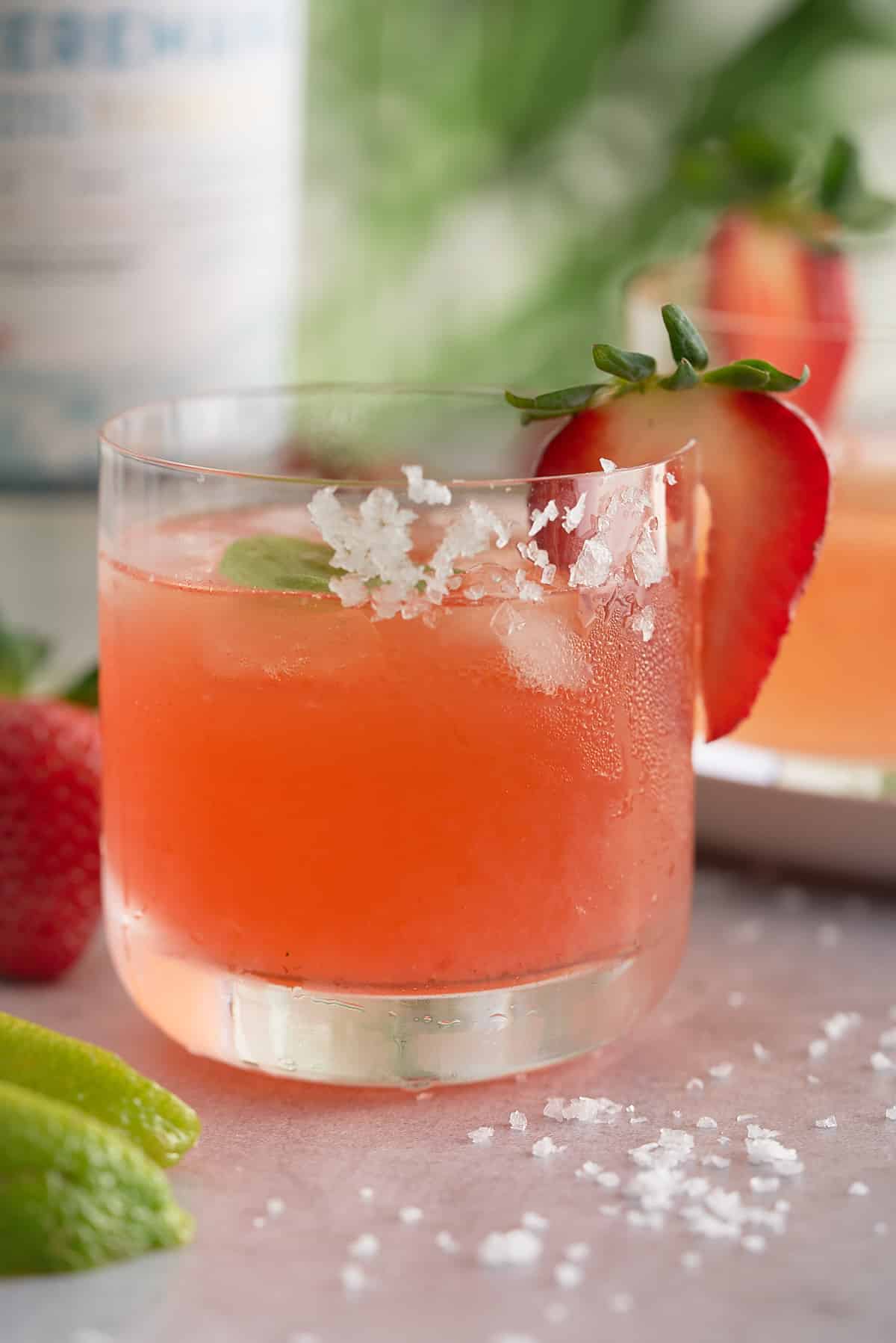 A glass filled with strawberry basil margarita served with ice and garnished with a slice of strawberry and a basil leaf.