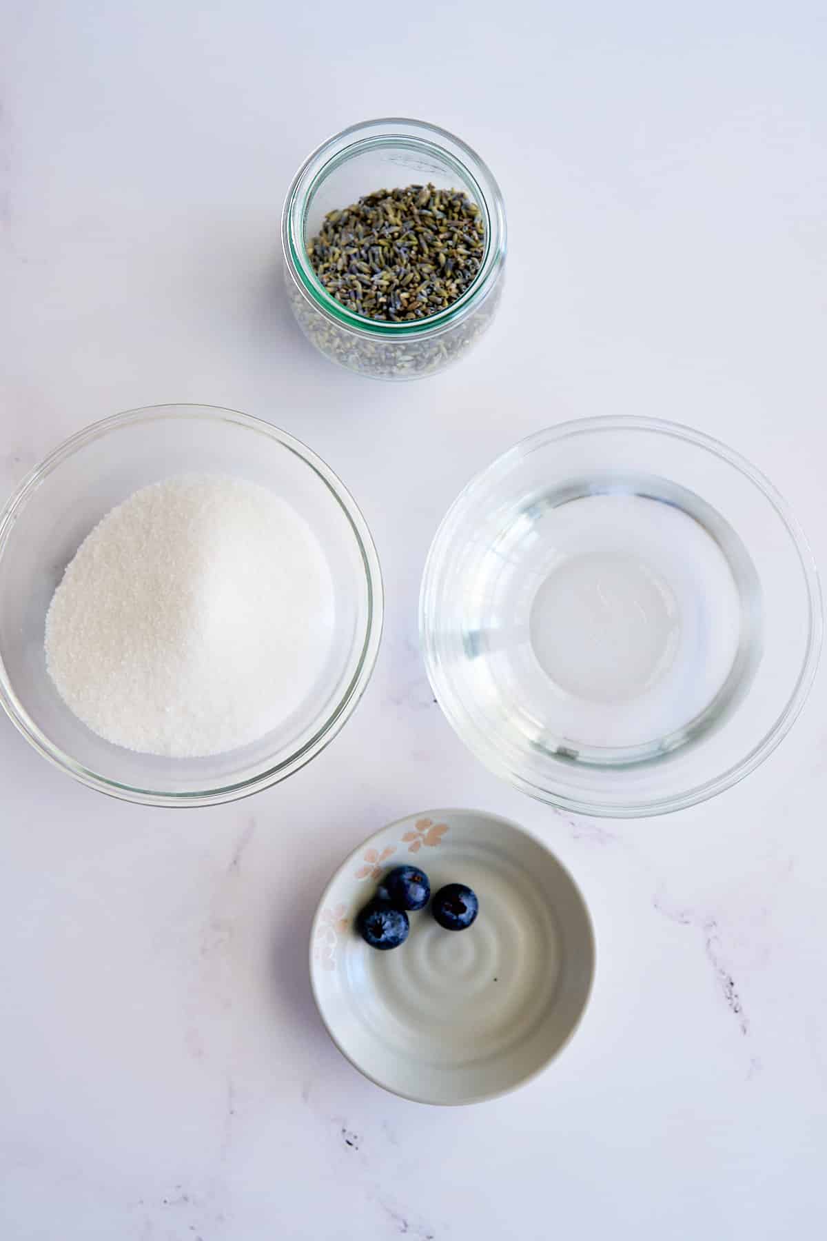 ingredients for the lavender syrup recipe on a white background. sugar, water, dried lavender leaves, and blueberries