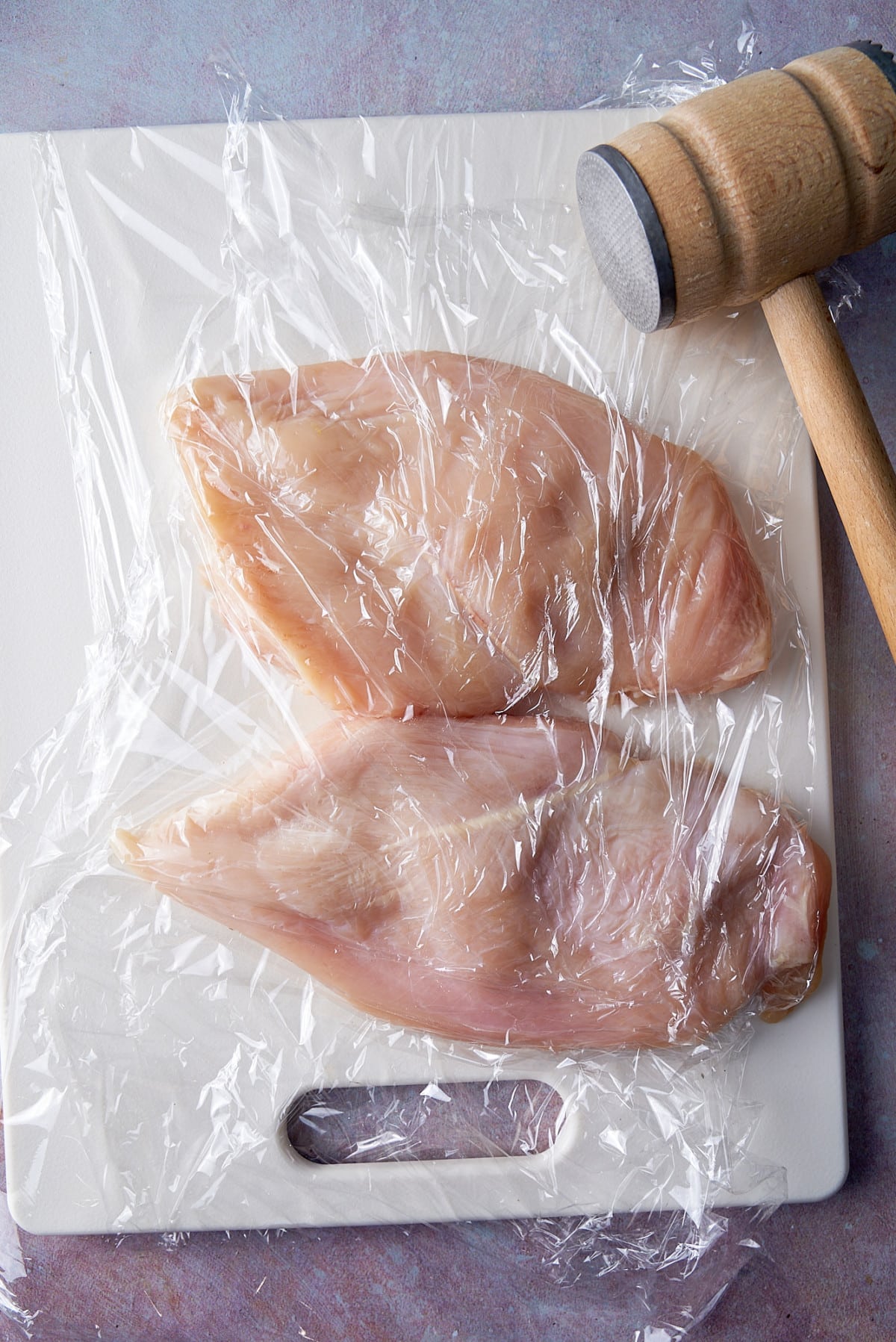 chicken breast under plastic wrap with meat cleaver next to it