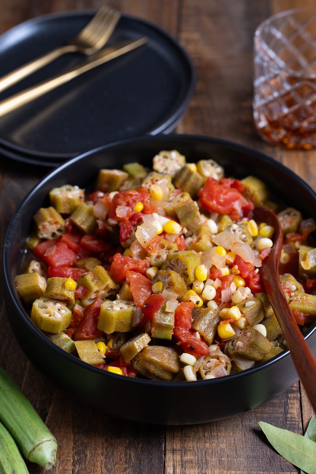 stewed okra and tomatoes in black bowl with plates in background and drink in background