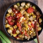 stewed okra and tomatoes with spoon in black bowl and fresh okra pods on the side of the dish