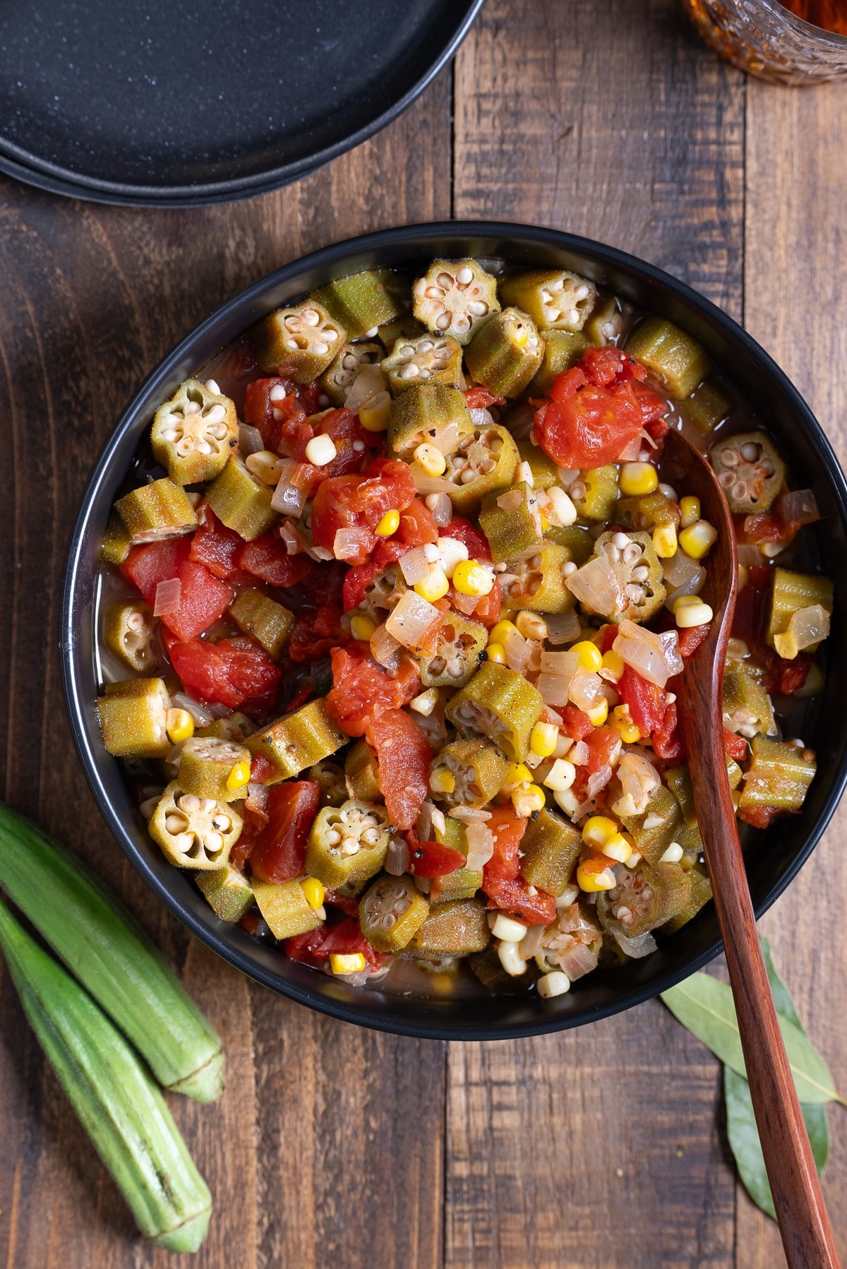 stewed okra and tomatoes with spoon in black bowl and fresh okra pods on the side of the dish