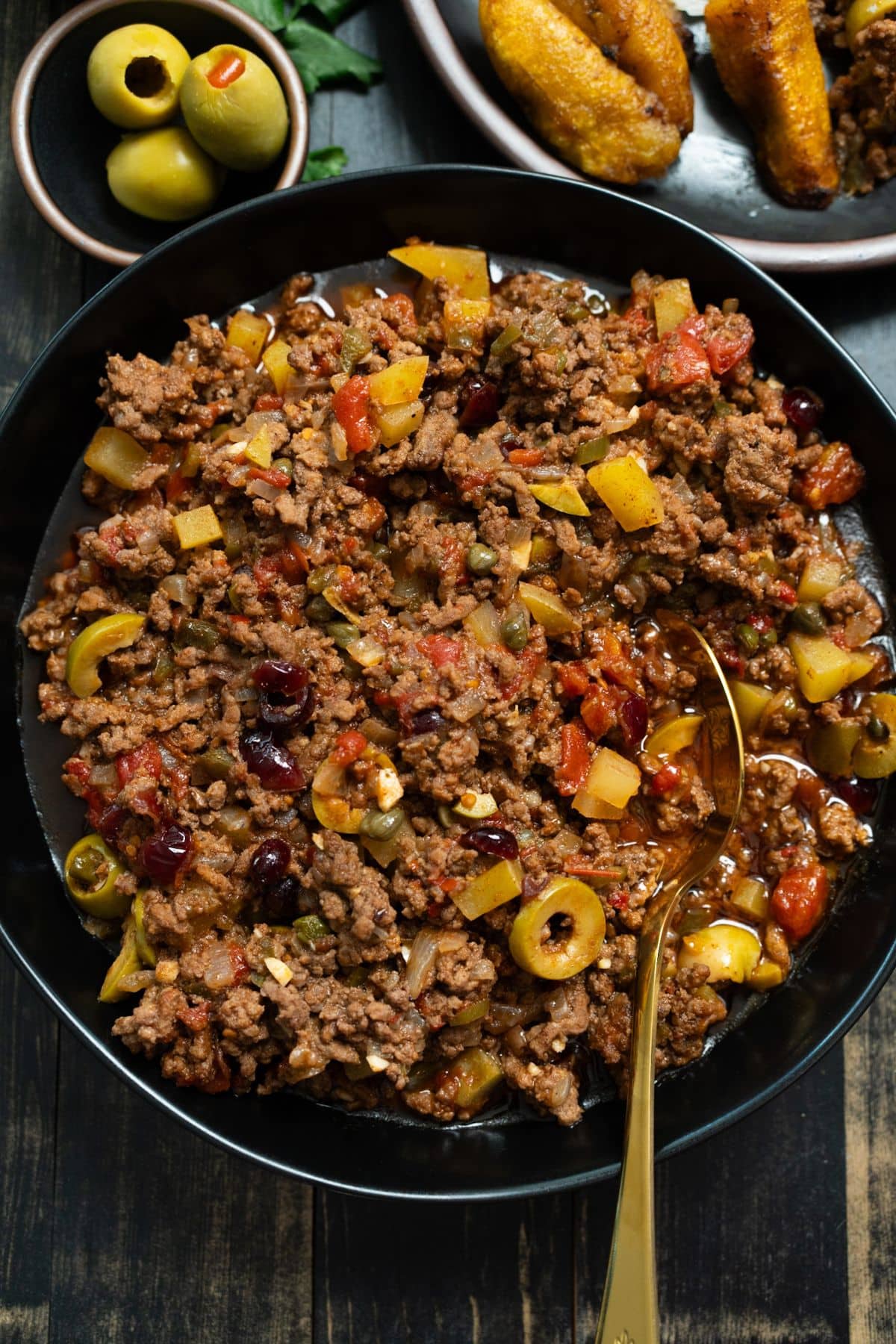Cuban picadillo and black bowl with gold spoon sticking out and olives on the side