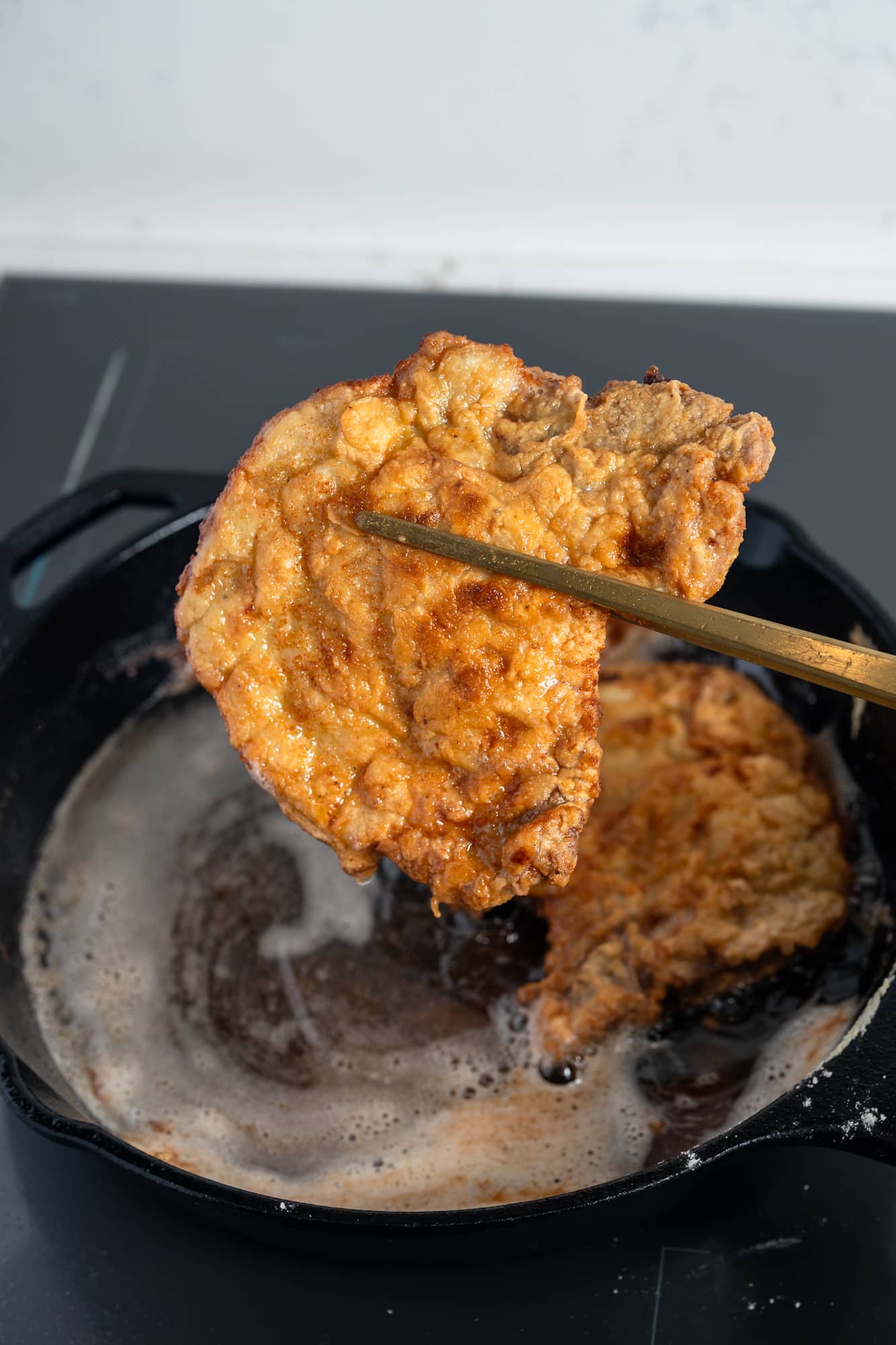 Lifting a crispy fried pork chop from a cast iron skillet with tongs.