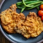 Two crispy fried pork chops served on a dark plate with fresh green beans and cherry tomatoes.