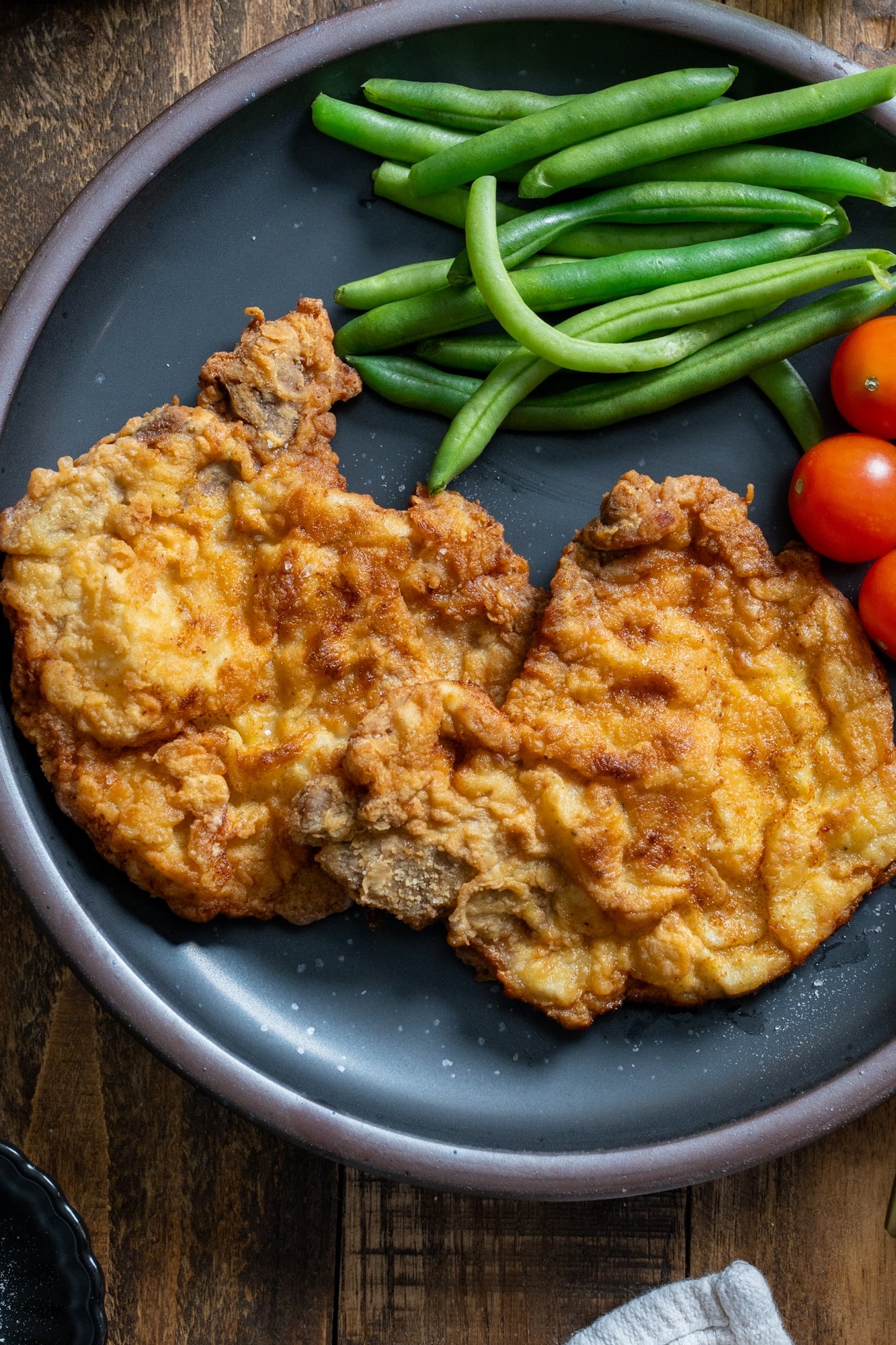Two crispy fried pork chops served on a dark plate with fresh green beans and cherry tomatoes.