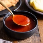 spoon lifting up sweet and sour sauce from a bowl with egg rolls and pineapple in the back