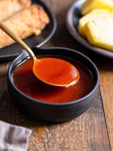 spoon lifting up sweet and sour sauce from a bowl with egg rolls and pineapple in the back