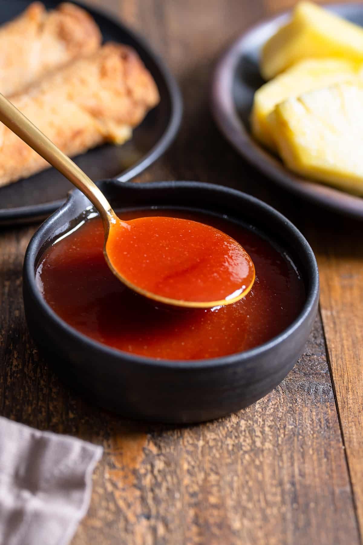 spoon lifting up sweet and sour sauce from a bowl with egg rolls and pineapple in the back