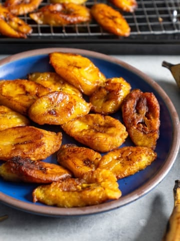 fried plantains on blue plate with plantains in rack in back