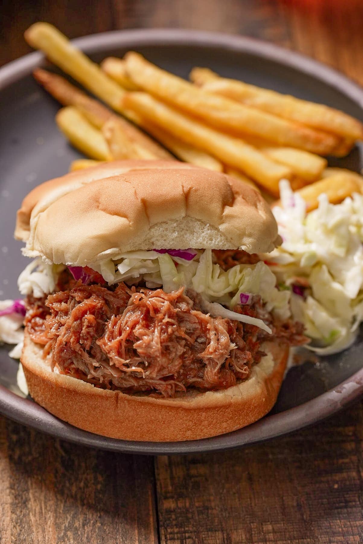 Pulled pork sandwich on bread with coleslaw and fries in the background.