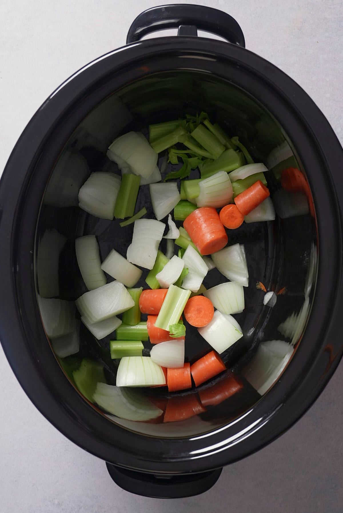 Chopped onion, celery, and carrots layered in the bottom of a black oval slow cooker insert.